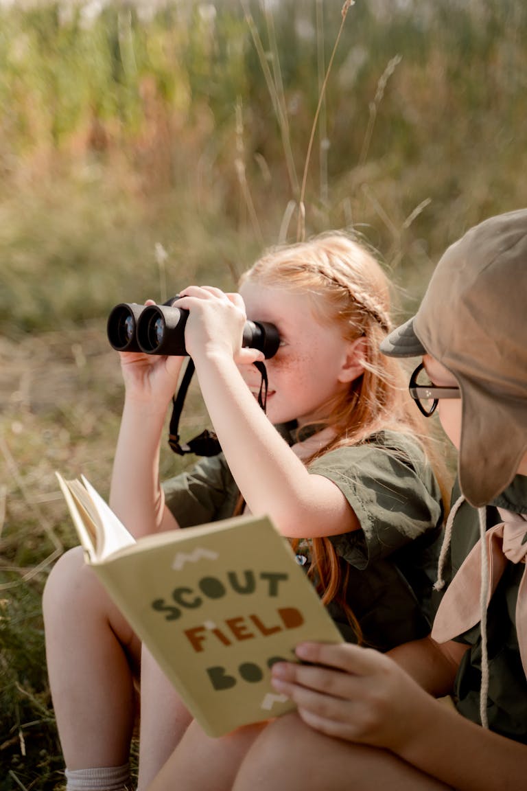 Two scouts sitting outdoors, one using binoculars, holding a scout field book.